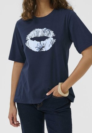 Femme portant un t-shirt bleu marine avec une grande empreinte de lèvres blanche, un jean foncé et un large bracelet beige à son poignet droit.
