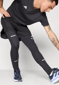 Shorts de sport noirs et leggings, avec des accents réfléchissants, associés à des chaussures de course bleu marine. Le matériau semble extensible et évacue l'humidité.