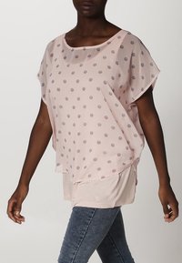 Blusa transparente rosa claro con lunares grises, corte holgado, escote amplio y mangas cortas. Superpuesta sobre una capa ajustada.
