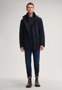 Cappotto navale con foderato puffer nero staccabile, chiusure con bottoni e zip, tasche laterali, abbinato a jeans blu e stivaletti marroni.