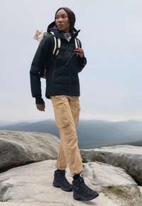Veste isolante noire avec capuche, haut gris clair, pantalon cargo beige et bottes de randonnée noires. Sacs à dos sur les épaules, à l'extérieur sur un terrain rocheux.