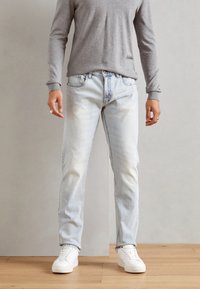 Jeans in denim azzurro chiaro con taglio dritto, finitura sbiadita e design a cinque tasche, abbinati a una camicia grigia a maniche lunghe e sneakers bianche.