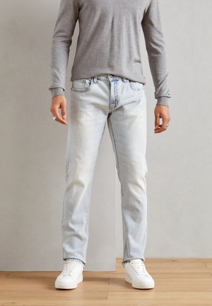 Jeans de mezclilla de un azul claro con un corte recto, acabado descolorido y diseño de cinco bolsillos, combinados con una camisa de manga larga gris y zapatillas blancas.