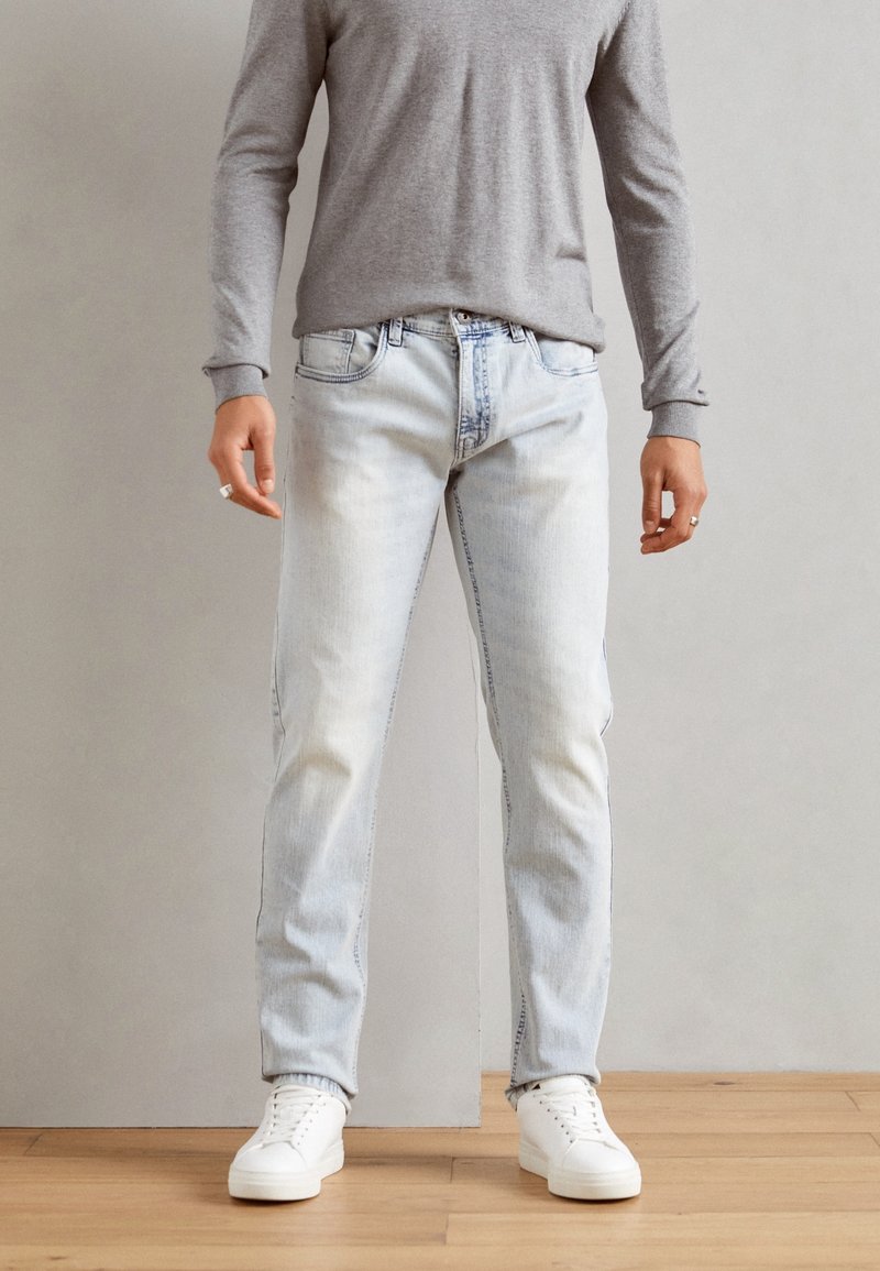 Jeans in denim azzurro chiaro con taglio dritto, finitura sbiadita e design a cinque tasche, abbinati a una camicia grigia a maniche lunghe e sneakers bianche.