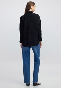Schwarzes Langarmshirt mit lockerer Passform und einer Rückenfalte. Kombiniert mit hellen, geraden Jeans und schwarzen, hohen Schuhen.