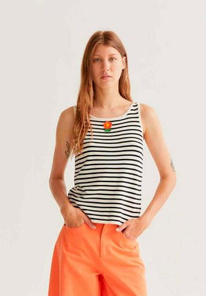 Joven mujer con top sin mangas a rayas negras y blancas con detalle de flor naranja, y pantalones de tiro alto de color naranja brillante, con las manos en los bolsillos.