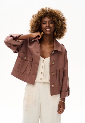 Kobieta z kręconymi włosami uśmiecha się, nosząc brązową zapinaną na guziki kurtkę na białym topie zapinanym na guziki oraz szerokie spodnie z subtelnym haftem.