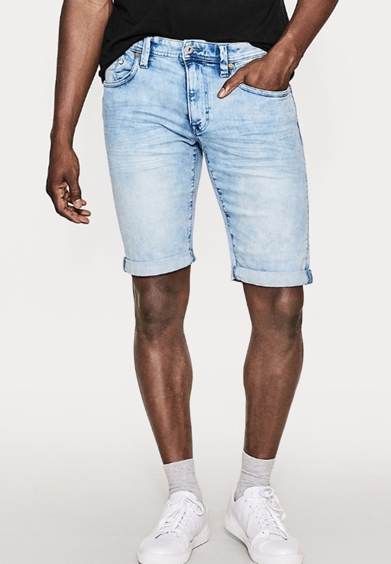 Shorts en denim bleu clair avec revers, cinq poches et une fermeture classique à boutons. Portés avec des baskets blanches et des chaussettes grises.