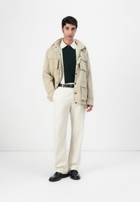 Doudoune beige clair avec plusieurs poches, portée par-dessus un polo vert foncé. Assortie à un pantalon crème et des chaussures noires.