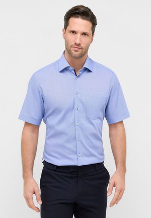 Mand iført en lyseblå kortærmet skjorte med brystlomme og mørke bukser, stående mod en ensfarvet lys baggrund.