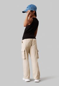 Top senza maniche nero, pantaloni cargo beige con tasche laterali e un cappellino in denim blu. Sneakers bianche completano il look.