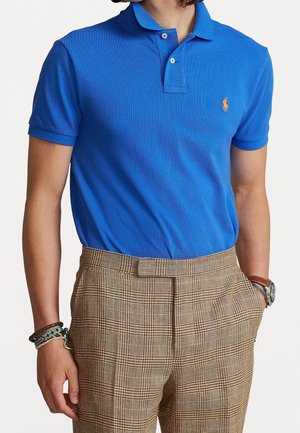 Personne portant un polo bleu à manches courtes et un pantalon à carreaux marron, avec des bracelets et une montre aux poignets.