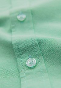 Tissu vert avec deux boutons translucides cousus sur une patte cousue, montrant la texture et les détails de couture fins.