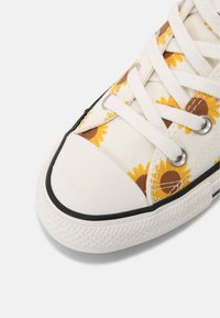 Zapatillas de lona blancas con estampado de girasoles. Presentan cordones blancos, ojales plateados, ribete de goma negra y una puntera texturizada.
