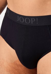 Gros plan sur une personne portant un slip noir avec une ceinture noire portant le nom de la marque "JOOP !" en lettres gris foncé.