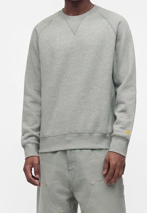Mann trägt hellgraues Sweatshirt mit Rundhalsausschnitt und passende Hose mit sichtbaren Nähten und goldenen Nieten an den Taschen.