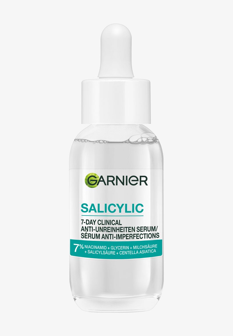 Bouteille en verre transparent avec un bouchon compte-gouttes blanc. L'étiquette présente un texte vert et turquoise. Contient un sérum transparent avec des bulles visibles.