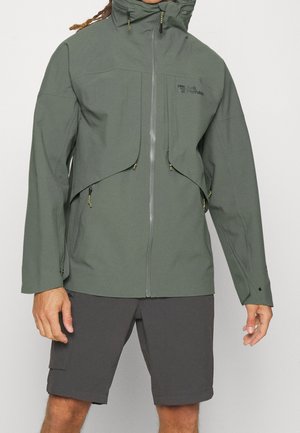 Mann trägt eine grüne Outdoor-Jacke mit Reißverschluss und dunkelgraue Shorts, steht vor einem schlichten hellen Hintergrund.