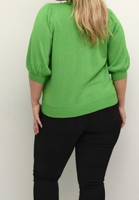 Pull en maille verte avec un motif texturé, manches bouffantes, et bords côtes au niveau des poignets et de l'ourlet, associé à un pantalon noir ajusté.