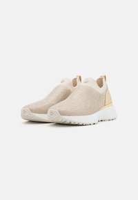 Beige slip-on sneakers med teksturert strikket overdel, gullaccenter på hælen, og en hvit såle med et mønster.