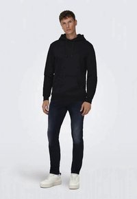 Svart hoodie med dragsnören, mjukt tyg, känguruficka, matchat med mörka jeans och vita sneakers, står på en ljus bakgrund.