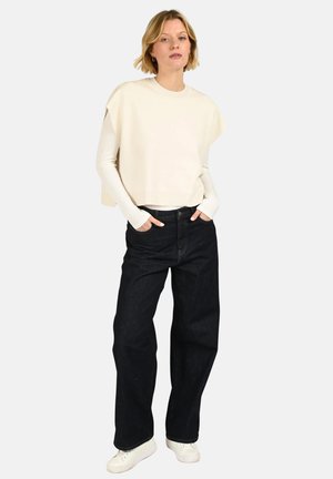 Personne blonde portant un haut crème sans manches par-dessus une chemise à manches longues, un jean noir à jambes larges et des baskets blanches, debout les mains dans les poches.