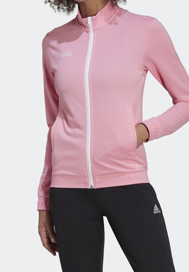 Chaqueta atlética de color rosa claro con cuello alto, cremallera frontal completa y dos bolsillos laterales. Presenta un logotipo blanco y un tejido suave y elástico.