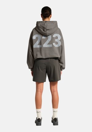 Person iført grå hættetrøje med stort "223" på ryggen, sorte shorts, hvide sokker med sort tekst og sorte sko, stående med ryggen mod.