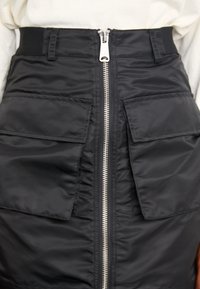 Black, padded skirt with a front zipper, featuring two large pockets and an elastic waistband. Material appears smooth with a slight sheen.