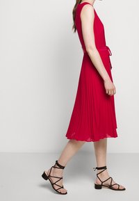 Červené plisované šaty s přiléhavým korzetem a páskem v oblasti pasu. Mají délku ke kolenům a jsou nošeny s černými sandály na řemínky.