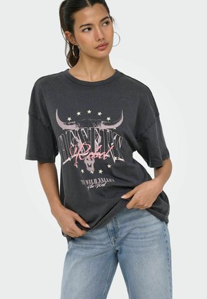 Ung kvinde med hoop-øreringe iført falmet sort oversized T-shirt med okseskalle og teksten "DESERT Rebel", kombineret med lyseblå jeans.