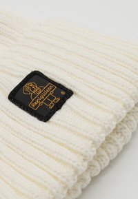 Gebreide witte muts met geribbelde textuur, voorzien van een zwart label met een oranje hondenafbeelding en merknaam, aan de voorkant vastgenaaid.