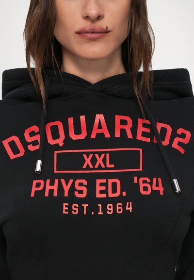 Donna che indossa un hoodie nero con testo rosso "DSQUARED2 XXL PHYS ED. '64 EST. 1964" e stringhe visibili su uno sfondo bianco.