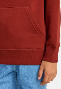 Felpa color bordeaux con polsini e orlo a costine, dotata di una tasca frontale. Abbinata a jeans in denim blu chiaro. Textura liscia.