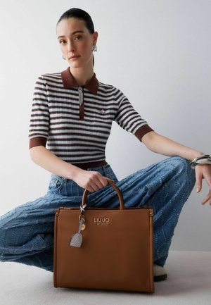 Ung kvinde sidder med benene krydset iført en stribet skjorte og teksturerede jeans, og holder en brun Liu Jo Milano håndtaske med nøglekæde.