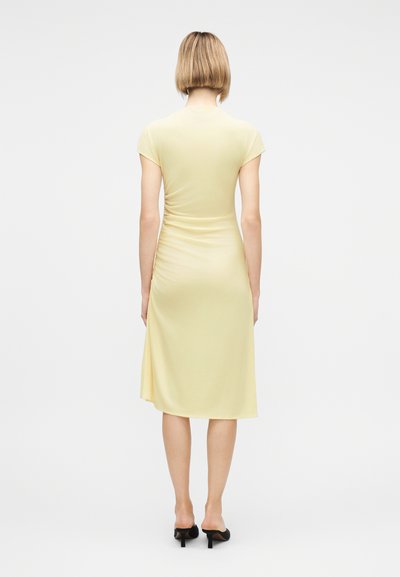 Γυναίκα όρθια που δείχνει την πίσω όψη ενός απαλά κίτρινου, μίντι φόρεμα με κοντά μανίκια και ρυτιδώσεις, φοράει μαύρα mule με τακούνι, σε μονόχρωμο φόντο.