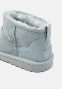 Botín de ante azul claro con forro suave, exterior texturizado, suela gris contrastante y acentos cosidos. Logo de UGG visible en la parte trasera.