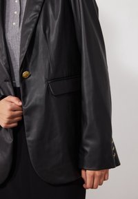 Blazer en cuir noir avec une coupe ajustée, fermeture à un bouton doré et une poche latérale. Le tissu présente une texture lisse et brillante.