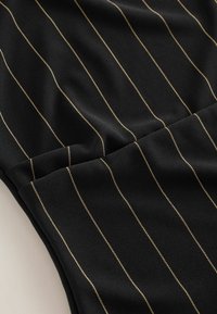 Tissu noir avec des rayures verticales dorées, présentant une texture lisse et un design plié. Les coutures sont visibles le long des lignes.