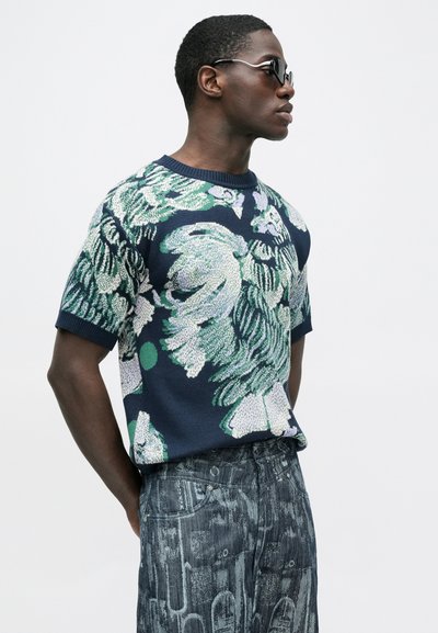 Μπλε σκούρο πουλόβερ με κοντά μανίκια, πράσινο λουλουδάτο σχέδιο, στρογγυλή λαιμόκοψη και ριμπ μανσέτες. Συνδυασμένο με γκρι τζιν παντελόνι με σχέδιο.