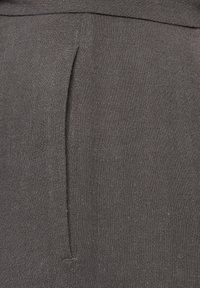 Close-up van grijze linnen stof met een verticale naad en plooidetail, waarbij de textuur en stiksels van het kledingstuk worden benadrukt.