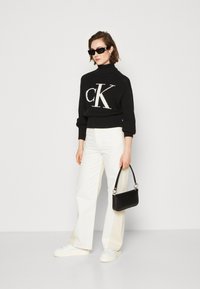 Ženska nosi črno pulover s turtleneck izrezom z velikim "ck" logotipom, bele široke hlače, bele superge, črno torbico in črne sončne očal.