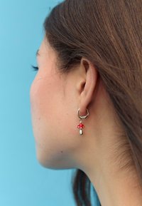Orecchino a cerchio in argento con un charm a fungo rosso, caratterizzato da una finitura lucida e dettagli rifiniti, indossato da un modello su uno sfondo blu.