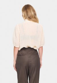 Blusa di raso crema con maniche a sbuffo, abbinata a pantaloni marroni a vita alta. La blusa ha una vestibilità ampia e un colletto rotondo.