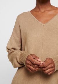 Personne portant un pull en tricot beige à col en V, les mains doucement croisées devant, avec des poignets côtelés visibles et des détails de couture.