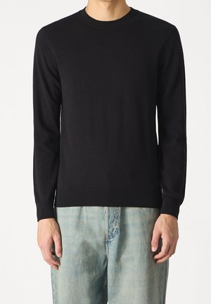 Man draagt een effen zwarte trui met ronde hals en lange mouwen, en lichtblauwe spijkerbroek tegen een effen achtergrond.