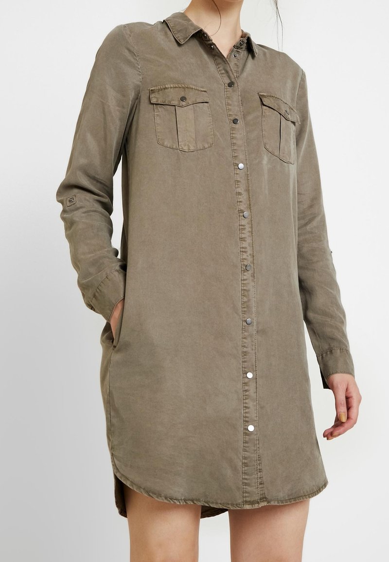 Robe-chemise olive à manches longues avec boutons-pression sur le devant, poches poitrine à rabat, col et poches latérales, portée avec une main dans la poche.