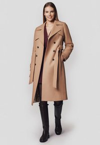 Płaszcz podwójnie zapinany w kolorze camel, wykonany z mieszanki wełny, z czarnymi guzikami, wyraźnymi klapami, paskiem w talii oraz bocznymi kieszeniami, noszony na ciemnej sukience i wysokich bucikach do kolan.