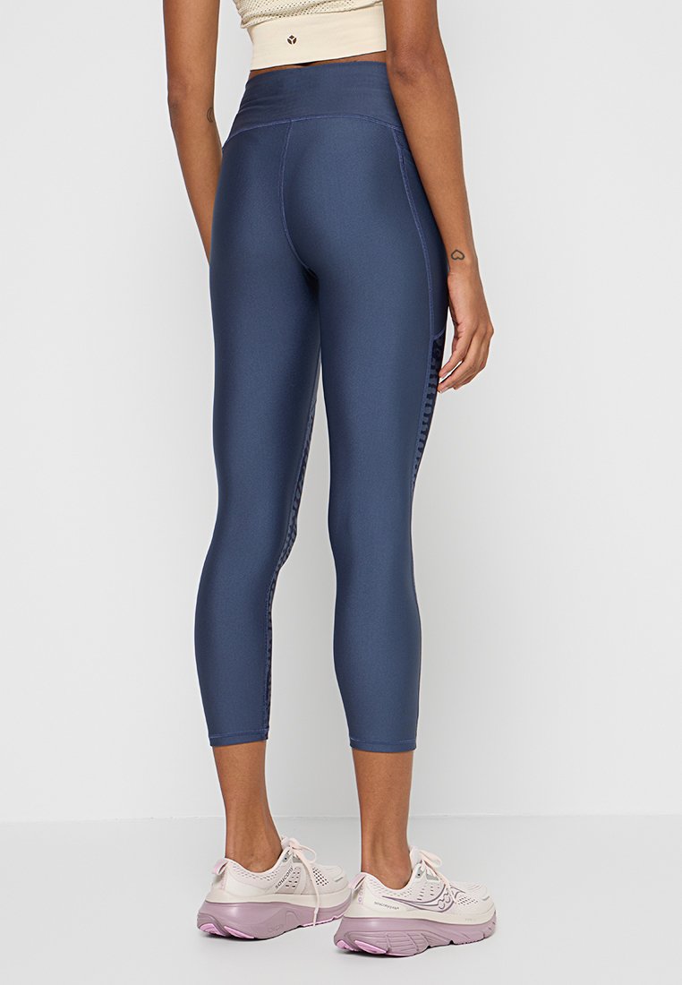 Mujer vestida con leggings cortos de talle alto azul marino y zapatillas de correr rosa claro, de pie contra un fondo claro y sencillo.