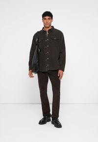 Camisa de pana marrón con dos bolsillos en el pecho, frente con botones, combinada con pantalones oscuros marrones y zapatos negros de cordones. Bolsa tote negra llevada al hombro.